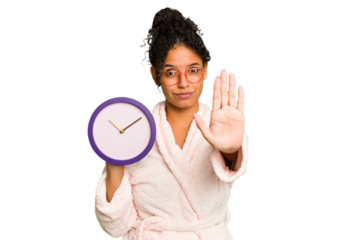 Young brazilian woman wearing a pajama holding a clock isolated standing with outstretched hand showing stop sign, preventing you.