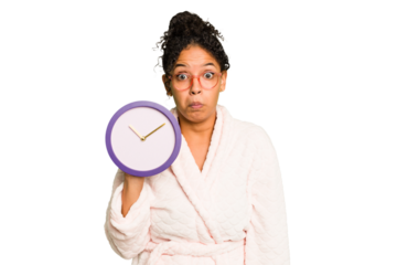 Young brazilian woman wearing a pajama holding a clock isolated shrugs shoulders and open eyes confused.