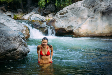 Fototapeta na wymiar Beautiful young woman wearing red bikini coming out stormy mountain river. Happy female brunette with sexy body on picturesque natural background.