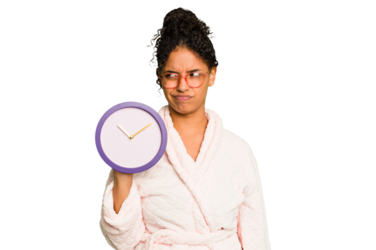 Young brazilian woman wearing a pajama holding a clock isolated confused, feels doubtful and unsure.