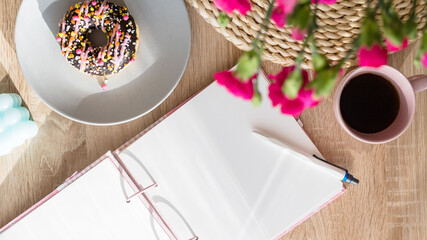 Cozy breakfast and planning. Notepad and pen, cup of coffee, pink donut, flowers and candles on the table. Women's desktop. Top view