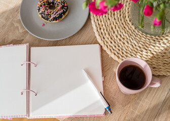 Cozy breakfast and planning. Notepad and pen, cup of coffee, pink donut, flowers and candles on the table. Women's desktop. Top view