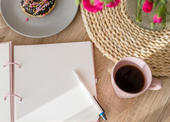 Cozy breakfast and planning. Notepad and pen, cup of coffee, pink donut, flowers and candles on the table. Women's desktop. Top view