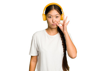 Young asian woman listening to music with headphones isolated with fingers on lips keeping a secret.