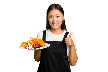 Young asian waitress woman holding waffles isolated smiling and raising thumb up