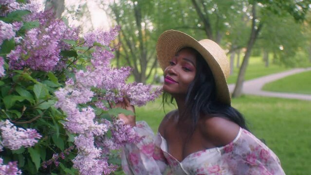 Black lady seductively smells touches pink Lilac flowers in park, eye contact