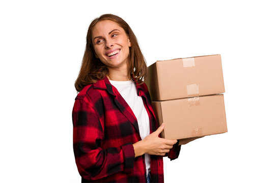 Young caucasian woman moving while picking up a box full of things isolated looks aside smiling, cheerful and pleasant.