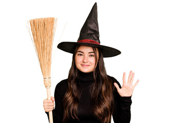 Young caucasian woman dressed as a witch holding a broom isolated on green chroma background smiling cheerful showing number five with fingers.