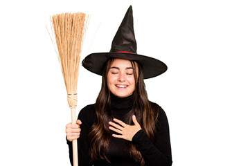 Young caucasian woman dressed as a witch holding a broom isolated on green chroma background laughs out loudly keeping hand on chest.