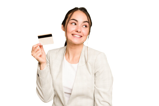 Young caucasian woman holding a credit card isolated on green chroma background looks aside smiling, cheerful and pleasant.