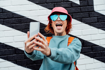Excited redhead woman screaming while taking a selfie photo outdoors. Emotional hipster fashion women in bright clothes, heart shaped glasses, bucket hat taking selfie photo on the phone camera - obrazy, fototapety, plakaty