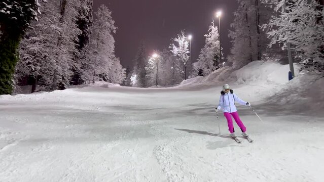 Active female skier enjoy evening skiing on snow slope path surrounded by fabulous forest where frost is on branches of trees. Illumination slope in mountains at ski resort at winter. Shot in movement