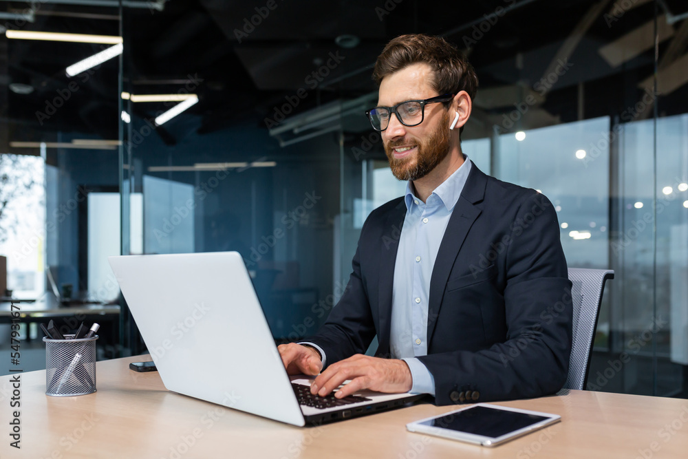 Wall mural Mature businessman in headphones small earbuds talking on a video call using a laptop, boss at work at the desk in a business suit in the middle of the office smiling friendly.