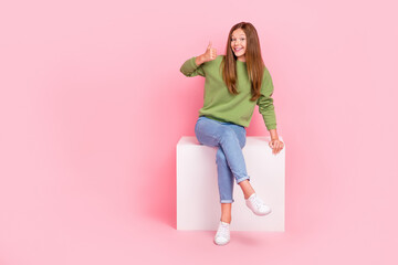 Full length photo of charming cute schoolgirl wear green sweatshirt sitting white chair thumb up empty space isolated pink color background