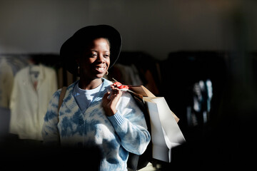 Trendy portrait of smiling black woman holding shopping bags in thrift store lit by sunlight, copy space