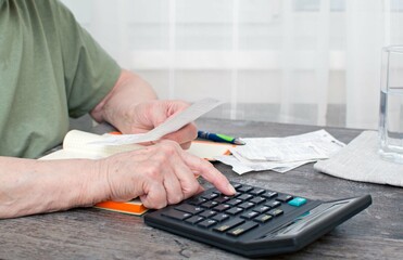 Fototapeta premium An elderly woman sitting at a table looks at receipts, bills from the store and counts on a calculator. Selective focus.