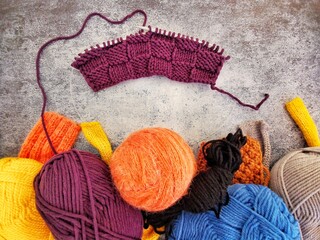 Purple piece of incomplete knitting with multicolored skeins of yarn on gray background top view. Knitted patterns and textures. Handmade and needlework hobbies. 