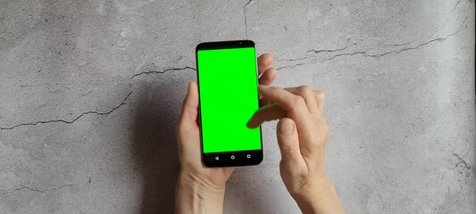 Old woman hand with smartphone with blank green screen simulating social network browsing or web surfing on white concrete background. Smartphone with copy space.