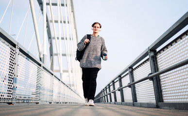 Confident young woman listening music with her headohones while walking across the footbridge. 