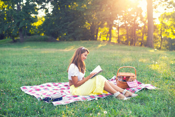 Young smiling woman sitting on meadow during picnic and reading book on a sunny summer day