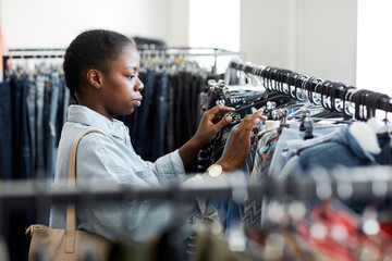 Side view portrait of black young woman looking at clothes on rack in second hand store
