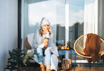 Smiling woman with blonde hair in winter clothes with coffe cup sitting near country house in winter