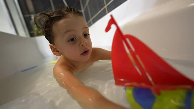 One little boy playing at bathtub with toy boat. Playful child bathing inside tub