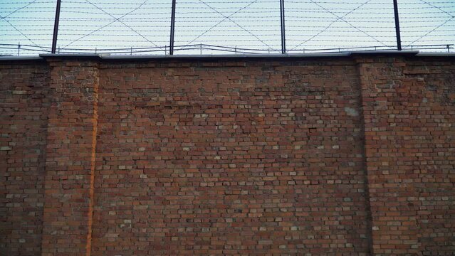 Low-angle view of jail prison dark red brick wall with a barbed wire fence on top with the cloudy sky, sad, gloomy, depressed atmosphere, tilt down shot