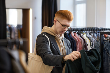 Young red haired man browsing clothes on racks while shopping sustainably in thrift shop