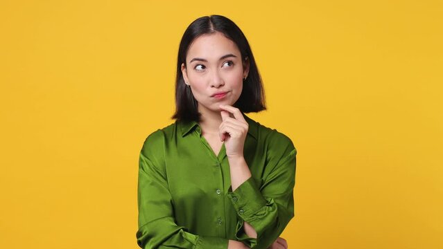 Excited pensive young woman of Asian ethnicity 20s she wear green shirt looks around think scratches at temple comes up with ideas raised finger get plan isolated on plain yellow color wall background