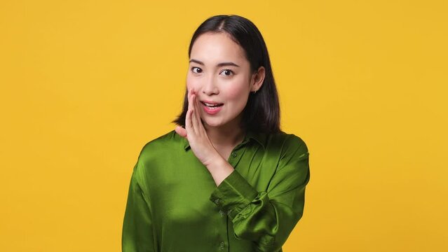 Mysterious funny young woman of Asian ethnicity 20s she wear green shirt whispering gossip and tells secret behind her hand sharing news isolated on plain yellow color wall background studio portrait