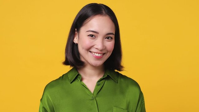 Beautiful toothy smiling charming friendly young woman of Asian ethnicity 20s she wears satin green shirt looking camera wink eye blink isolated on plain yellow color wall background studio portrait