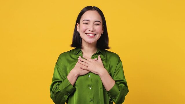 Smiling surprised good kind happy young woman of Asian ethnicity 20s she wear satin green shirt ask who for me oh it so sweet put hands on chest isolated on plain yellow color wall background studio