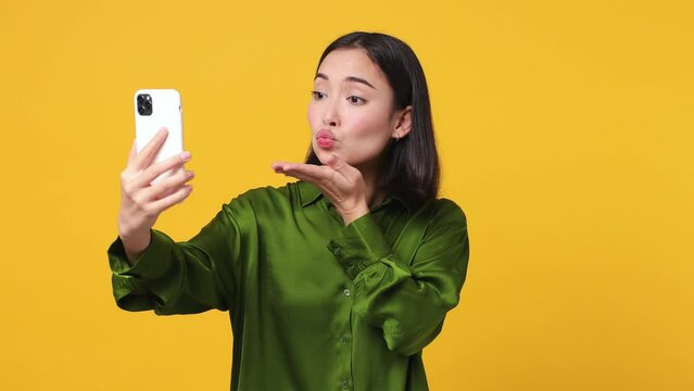 Beautiful young woman of Asian ethnicity she wear green shirt posing doing selfie shot on mobile cell phone post photo on social network isolated on plain yellow color wall background studio portrait