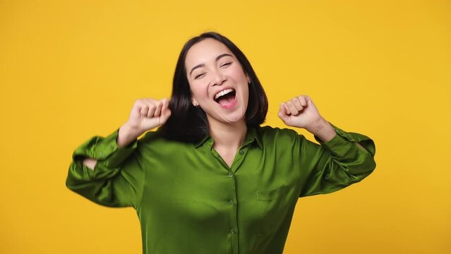 Beautiful fancy vivid happy bright young woman of Asian ethnicity she wear green shirt dance rising have fun enjoy relax celebrate play fluttering hair isolated on plain yellow color wall background