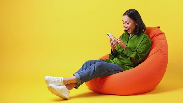 Full size body length smiling young woman wear green shirt sit in bag chair hold use point finger on mobile cell phone just found out great good news isolated on plain yellow color background studio