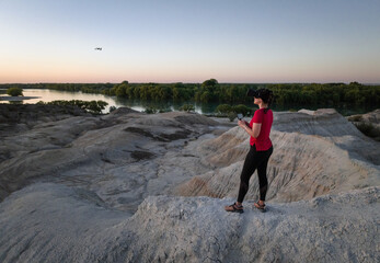Young woman flying FPV drone with Virtual reality mask in semi-desert area by the river.