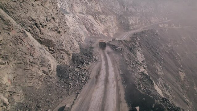 Aerial view of full-loaded tipper fastly rushing on dusty road in a huge quarry. Back view of yellow mining truck driving across the work site in opencast mine