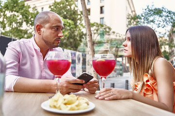 Side view of loving couple sitting at table with cocktails in glasses and looking at each other while enjoying date in patio of summer cafe