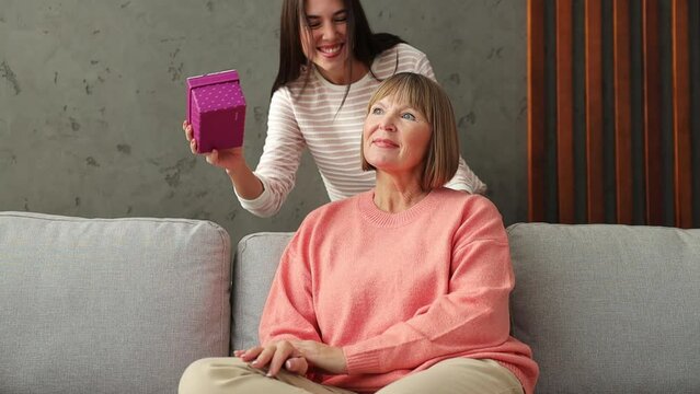 Two adult women older mature mom young kid receiving gift box hugs kiss sits on gray sofa couch stay home flat rest relax spend free spare time in living room indoor. Mother's Day love family concept