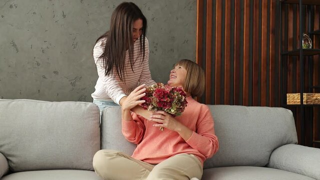 Two adult women older mature mom young kid receiving flowers hugs kiss sits on gray sofa couch stay home flat rest relax spend free spare time in living room indoor. Mother's Day love family concept