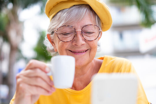 Smiling Senior Woman Having Relaxed Moment With An Espresso Coffee Cup At Outdoor Cafe Looking At Mobile Phone. Attractive Caucasian Elderly Lady In Yellow Holding White Coffee Cup