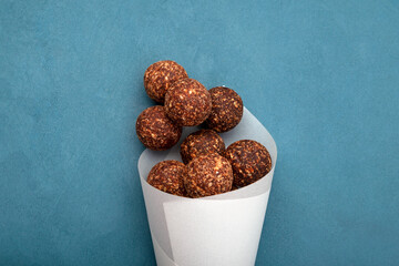 Energy balls with dried date fruits and carob powder. Vegetarian round candies in paper packaging on blue textured background, selective focus. Raw vegan alternative sweets