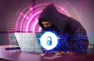 A male spy hacker in a black hoodie sits on a table in a dark room. looking at laptop computer