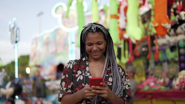 Video of an African American young woman taking selfies happily at amusement park during music festival, checking photo and posting on social media apps. Girl using mobile to make pictures outdoors.