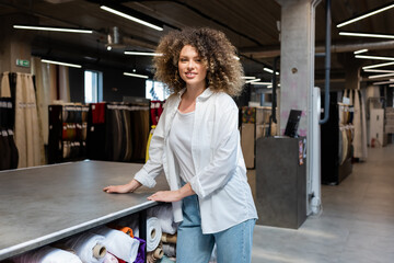 cheerful and curly saleswoman standing near rack with different fabric rolls in textile shop