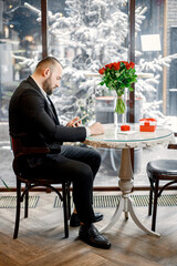 Fototapeta premium Man sitting in restaurant and waiting for his girlfriend with bouquet of red roses