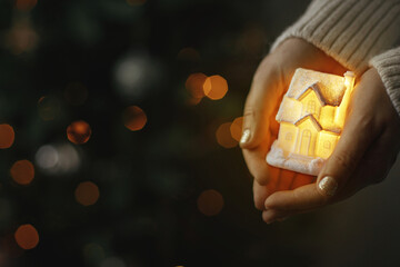 Hands holding illuminated little house on background of christmas tree with lights bokeh. Cozy...