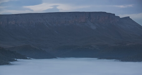 Mountain evening landscape with low clouds in the valley and a steep cliff in the background, panorama of the evening mountains