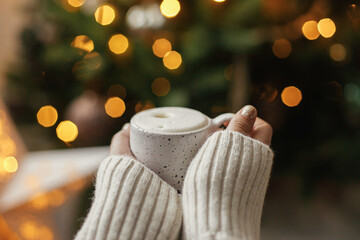Atmospheric winter hygge. Hands holding warm cup of coffee on background of christmas tree with lights. Cozy home. Woman hands in warm sweater holding  stylish mug at illumination bokeh